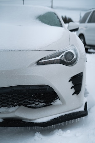 A white car is covered in ice and snow, with icicles hanging from its front bumper. The headlight and front grille are partially visible through the frost. Another vehicle is parked nearby, also coated in snow.