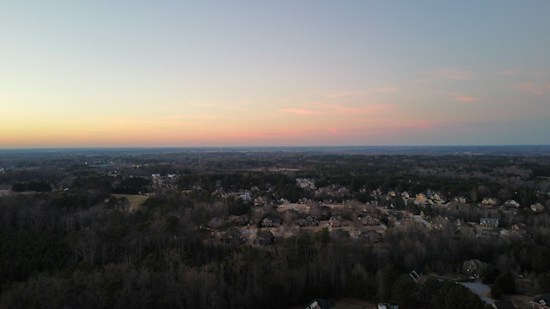 A panoramic view of a residential area surrounded by dense forests under a serene evening sky. The horizon glows with soft pastel hues of orange, pink, and blue, blending into the clear sky above. The houses are nestled closely together, displaying a suburban landscape.