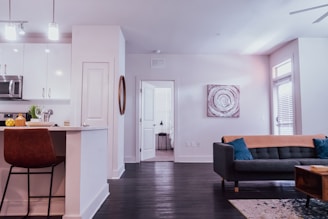 A modern living space featuring a sleek kitchen with white cabinetry and stainless steel appliances on the left. A countertop with a brown leather chair, decorative items, and a plant add warmth. Towards the right, a cozy living area with a dark grey sofa adorned with blue and orange cushions is visible. A textured canvas artwork hangs above. Wooden flooring and neutral walls create an inviting and comfortable atmosphere. Natural light streams in through a window near the sofa, adding to the bright ambiance.