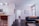 A modern living space featuring a sleek kitchen with white cabinetry and stainless steel appliances on the left. A countertop with a brown leather chair, decorative items, and a plant add warmth. Towards the right, a cozy living area with a dark grey sofa adorned with blue and orange cushions is visible. A textured canvas artwork hangs above. Wooden flooring and neutral walls create an inviting and comfortable atmosphere. Natural light streams in through a window near the sofa, adding to the bright ambiance.
