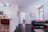 A modern living space featuring a sleek kitchen with white cabinetry and stainless steel appliances on the left. A countertop with a brown leather chair, decorative items, and a plant add warmth. Towards the right, a cozy living area with a dark grey sofa adorned with blue and orange cushions is visible. A textured canvas artwork hangs above. Wooden flooring and neutral walls create an inviting and comfortable atmosphere. Natural light streams in through a window near the sofa, adding to the bright ambiance.