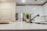 A modern kitchen with a sleek design features white cabinets and a stainless steel refrigerator. The focus is on a marble countertop with a silver faucet and a potted plant. Two wine glasses are placed on plates, adding to the minimalist aesthetic.