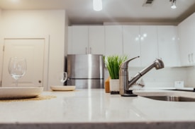 A modern kitchen with a sleek design features white cabinets and a stainless steel refrigerator. The focus is on a marble countertop with a silver faucet and a potted plant. Two wine glasses are placed on plates, adding to the minimalist aesthetic.