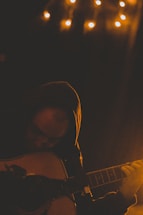 A person wearing a hoodie playing an acoustic guitar in a dimly lit room. Small warm lights hang above, creating a cozy and intimate atmosphere. Shadows and low lighting emphasize the contemplative mood.