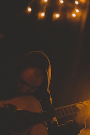 A person wearing a hoodie playing an acoustic guitar in a dimly lit room. Small warm lights hang above, creating a cozy and intimate atmosphere. Shadows and low lighting emphasize the contemplative mood.
