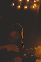 A person wearing a hoodie playing an acoustic guitar in a dimly lit room. Small warm lights hang above, creating a cozy and intimate atmosphere. Shadows and low lighting emphasize the contemplative mood.