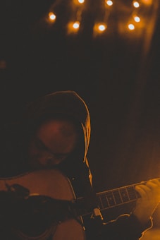 A person wearing a hoodie playing an acoustic guitar in a dimly lit room. Small warm lights hang above, creating a cozy and intimate atmosphere. Shadows and low lighting emphasize the contemplative mood.