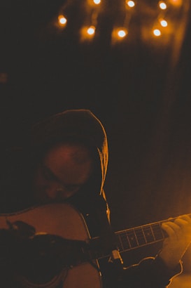 A person wearing a hoodie playing an acoustic guitar in a dimly lit room. Small warm lights hang above, creating a cozy and intimate atmosphere. Shadows and low lighting emphasize the contemplative mood.