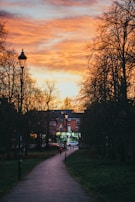 A pathway in a park lined with bare trees on both sides, leading towards a building with soft lighting in the windows. The sky above is an intense mix of orange and yellow hues typical of a sunset. A streetlamp stands alongside the path, and a small figure in a red jacket can be seen walking towards the building.