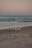 A group of four surfers carrying surfboards walks along a beach during sunset. The sky is painted with soft pastel colors, and the ocean waves gently lap against the shore. A solitary person can be seen in the distance near the water.