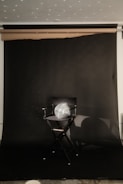 A director's chair is placed against a dark backdrop with a reflective disco ball resting on the seat. Light reflects off the disco ball, creating scattered spots of light on the backdrop. The setting appears to be a studio or a staged area for photography or film.