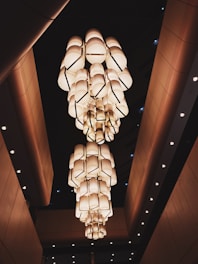 A series of large, modern chandeliers hang from a high ceiling. The chandeliers are composed of numerous cylindrical light fixtures, creating an impressive and geometric visual pattern. The environment appears to be an upscale indoor space with sleek architectural elements.