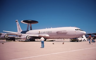 A military aircraft displayed on a runway, marked with 'U.S. AIR FORCE' on its fuselage. It has a large rotating radar dome on top and is surrounded by several people observing or walking by. The sky is clear and blue, and there are no clouds visible.