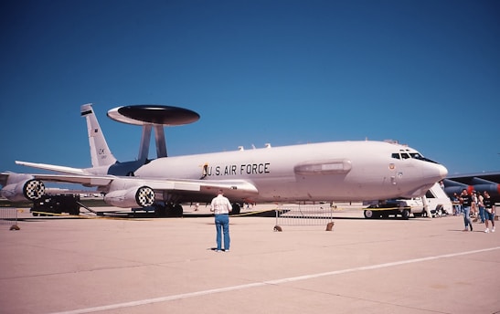 A military aircraft displayed on a runway, marked with 'U.S. AIR FORCE' on its fuselage. It has a large rotating radar dome on top and is surrounded by several people observing or walking by. The sky is clear and blue, and there are no clouds visible.