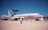 A military aircraft displayed on a runway, marked with 'U.S. AIR FORCE' on its fuselage. It has a large rotating radar dome on top and is surrounded by several people observing or walking by. The sky is clear and blue, and there are no clouds visible.
