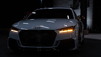 A sleek, modern white car with illuminated headlights is prominently showcased against a dimly lit urban background. The vehicle has a distinctive grille and sharp, angular design lines, providing a sense of sophistication and power.