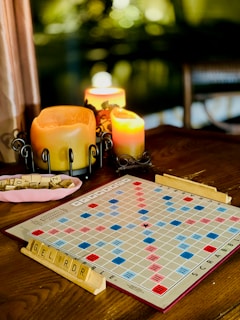 A cozy setting with a Scrabble board on a wooden table. Letter tiles are placed on racks, while several lit candles provide a warm glow. The background has a dim, blurred appearance, creating a serene ambiance.