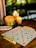 A cozy setting with a Scrabble board on a wooden table. Letter tiles are placed on racks, while several lit candles provide a warm glow. The background has a dim, blurred appearance, creating a serene ambiance.