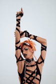 A person with orange hair styled in an avant-garde manner, wearing a black outfit with intricate strap designs. The outfit includes a choker with metal rings and long gloves. The makeup is bold, featuring dark lipstick and dramatic eyeliner. The background is plain and light-colored.