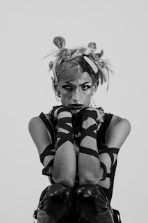 A person with a unique hairstyle featuring knots and hair clips is seated in a crouched position. They have dramatic makeup and are dressed in a sleeveless top with crisscrossed bands wrapping around their arms. The overall style is edgy and expressive.