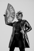 A person dressed in a dramatic outfit, wearing a long, shiny leather jacket and holding an ornate fan with Asian-style patterns. Their makeup is bold, with striking facial features, particularly focusing on the eyes and lips. The hair is styled in an edgy fashion, with a small horn accessory on the forehead.