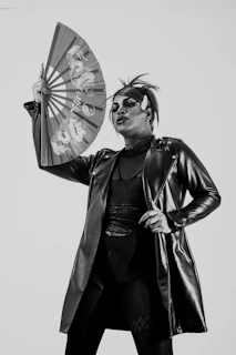 A person dressed in a dramatic outfit, wearing a long, shiny leather jacket and holding an ornate fan with Asian-style patterns. Their makeup is bold, with striking facial features, particularly focusing on the eyes and lips. The hair is styled in an edgy fashion, with a small horn accessory on the forehead.