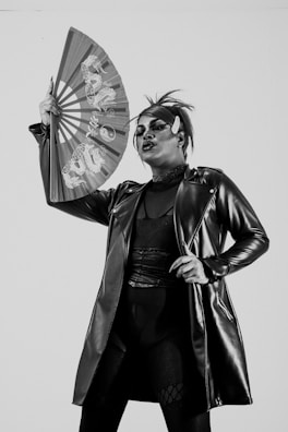 A person dressed in a dramatic outfit, wearing a long, shiny leather jacket and holding an ornate fan with Asian-style patterns. Their makeup is bold, with striking facial features, particularly focusing on the eyes and lips. The hair is styled in an edgy fashion, with a small horn accessory on the forehead.