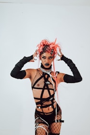 A person wearing a bold, avant-garde outfit with fishnet and straps, accessorized with arm-length gloves. The individual's pink hair is styled in an eclectic, dramatic fashion, and their makeup is dark and striking, featuring black lipstick.