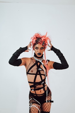A person wearing a bold, avant-garde outfit with fishnet and straps, accessorized with arm-length gloves. The individual's pink hair is styled in an eclectic, dramatic fashion, and their makeup is dark and striking, featuring black lipstick.