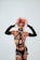 A person wearing a bold, avant-garde outfit with fishnet and straps, accessorized with arm-length gloves. The individual's pink hair is styled in an eclectic, dramatic fashion, and their makeup is dark and striking, featuring black lipstick.