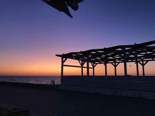 The image captures a seaside at dusk with a vibrant gradient in the sky, transitioning from deep blue to orange near the horizon. A wooden structure with a slatted roof is silhouetted against the colorful backdrop. The calm sea spreads out along the horizon, enhancing the tranquil scene.