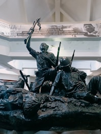 A bronze statue depicting a group of soldiers in a dynamic pose, with one figure prominently raising an object in triumph. The figures are dressed in military attire, and the sculpture captures a moment of action and struggle. Above the statue, there's a banner with text and a photograph.