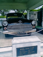 A vintage black car, identified as an Opel Kapitan from 1956, is displayed indoors. The car is roped off, and an informational plaque is prominently placed in front, detailing the vehicle's make and history. The setting is well-lit and appears to be a museum or exhibit space.