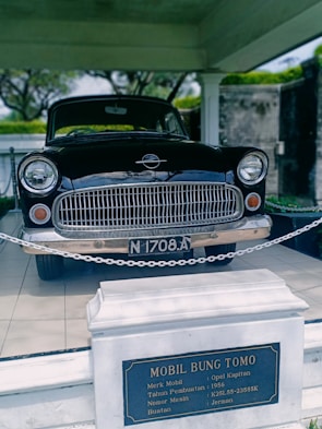 A vintage black car, identified as an Opel Kapitan from 1956, is displayed indoors. The car is roped off, and an informational plaque is prominently placed in front, detailing the vehicle's make and history. The setting is well-lit and appears to be a museum or exhibit space.