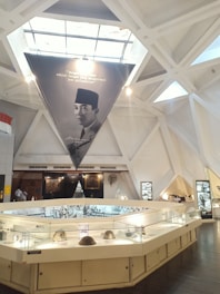 An exhibition space with a modern architectural ceiling featuring triangular shapes and skylights. A large triangular banner with a portrait of a person wearing traditional attire hangs prominently. Below the banner, an illuminated display case holds several objects, including what appear to be helmets and historical artifacts. The walls display photographs and other framed items.