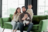 A family of four is seated on a green couch. A woman and a man are sitting with their children, one of whom is a baby held by the man. The other child is standing on the couch next to them. They are dressed in cozy, neutral-toned clothing, and appear to be in a bright, modern room with large windows.