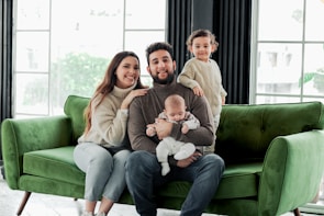 A family of four is seated on a green couch. A woman and a man are sitting with their children, one of whom is a baby held by the man. The other child is standing on the couch next to them. They are dressed in cozy, neutral-toned clothing, and appear to be in a bright, modern room with large windows.