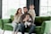A family of four is seated on a green couch. A woman and a man are sitting with their children, one of whom is a baby held by the man. The other child is standing on the couch next to them. They are dressed in cozy, neutral-toned clothing, and appear to be in a bright, modern room with large windows.