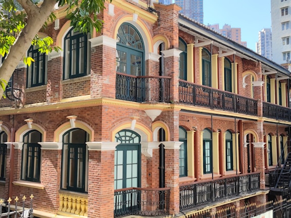 A historic building with classic red brick architecture and arched windows, featuring dark green frames and ornate black iron railings. The building's two stories include multiple windows that allow natural light to illuminate the facade. A tree with green leaves partially obstructs the view, adding a natural element to the urban setting.