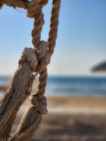 Thick braided ropes with visible knots are showcased in focus, against a blurry background of a beach and a blue sky. The ropes have a coarse texture and are hanging in a relaxed manner.