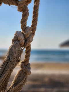 Thick braided ropes with visible knots are showcased in focus, against a blurry background of a beach and a blue sky. The ropes have a coarse texture and are hanging in a relaxed manner.