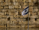A stone wall with large, ancient blocks is in the background. In the foreground, a flag with a blue Star of David flutters on a metal flagpole.