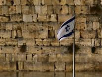 A stone wall with large, ancient blocks is in the background. In the foreground, a flag with a blue Star of David flutters on a metal flagpole.