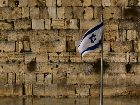 A stone wall with large, ancient blocks is in the background. In the foreground, a flag with a blue Star of David flutters on a metal flagpole.