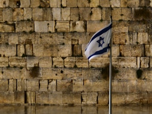 A stone wall with large, ancient blocks is in the background. In the foreground, a flag with a blue Star of David flutters on a metal flagpole.