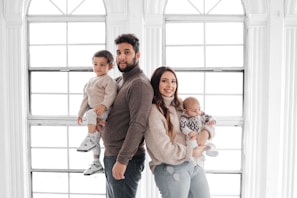 A family portrait featuring two adults and two children standing in front of large, bright windows. The father holds a toddler, while the mother holds a baby. They are all dressed in cozy, neutral-toned sweaters and jeans.