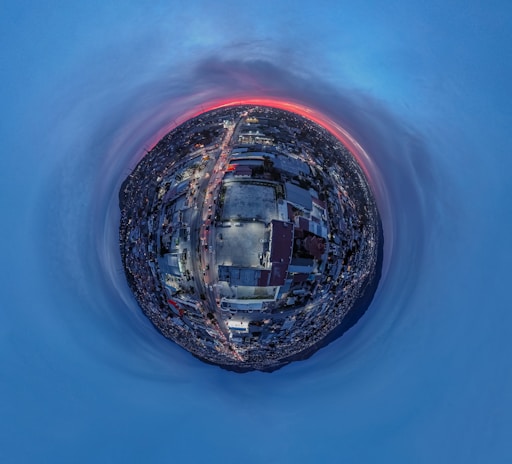 A spherical panorama capturing an urban landscape at dusk, with a prominent road leading through densely packed buildings. The sky is filled with shades of blue, transitioning to a vibrant orange and red horizon.