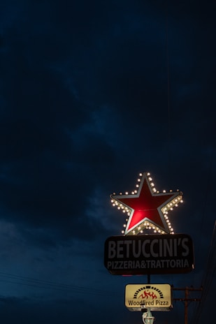 A brightly lit restaurant sign stands against a dark evening sky. The sign features a large red star outlined by small white lights. Below the star, there's text reading 'BETUCCINI'S PIZZERIA & TRATTORIA', followed by a lit panel advertising 'Woodfired Pizza' with a graphic depicting flames.
