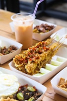 A variety of food items is displayed on a wooden table, featuring fried skewered chicken topped with crispy garnishes and slices of raw cucumber on the side. In the background, a plastic cup filled with ice and a pink straw containing a creamy iced beverage is visible.
