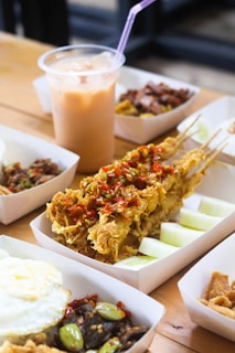 A variety of food items is displayed on a wooden table, featuring fried skewered chicken topped with crispy garnishes and slices of raw cucumber on the side. In the background, a plastic cup filled with ice and a pink straw containing a creamy iced beverage is visible.